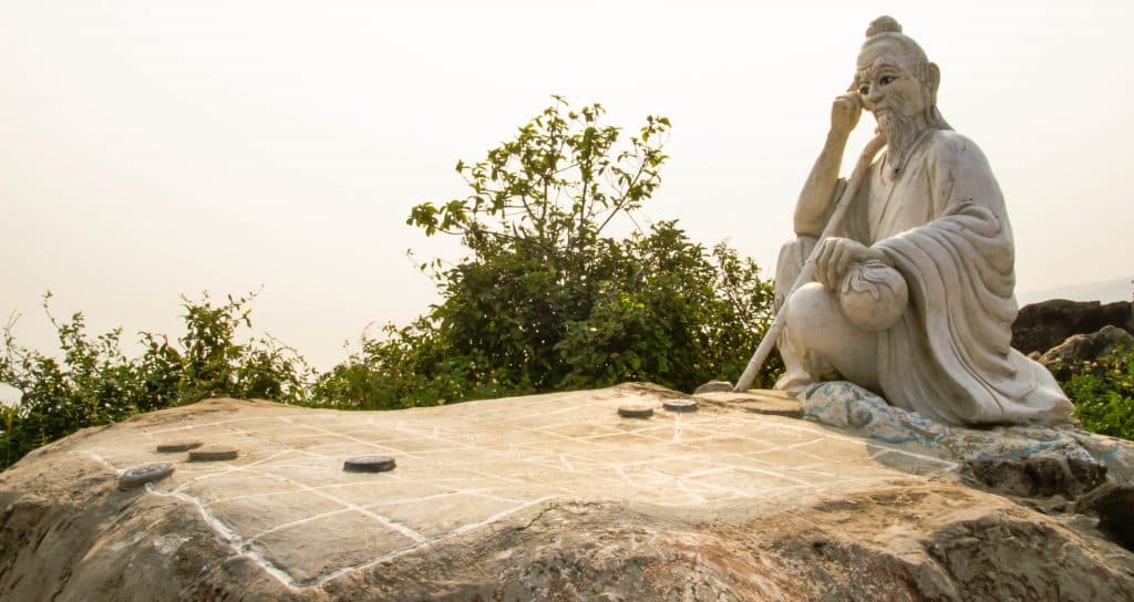 A statue of Confucius on a rock in Vietnam. The statue sits crosslegged over a chess board. 