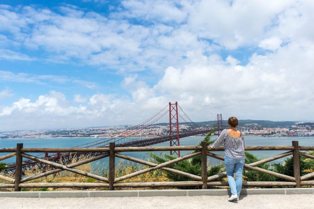 a girl dressed in black and white top and blue jeans stands with her back to the camera and looks at the 25th of april bridge in lisbon from a viewpoint in christo rei
