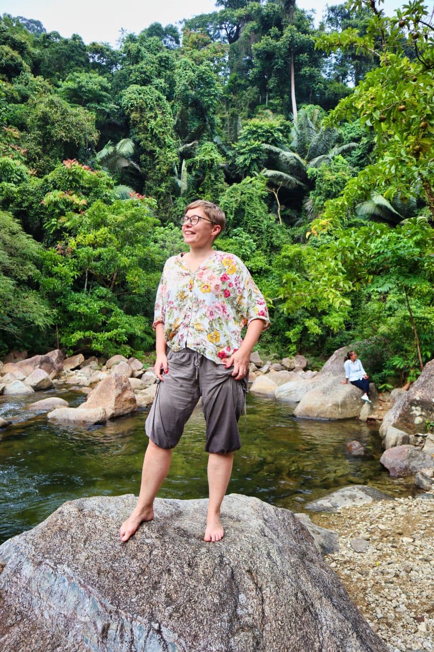 a woman dressed in brown trousers and a flowery shirt stands on a rock at the promlok waterfall smiling. 