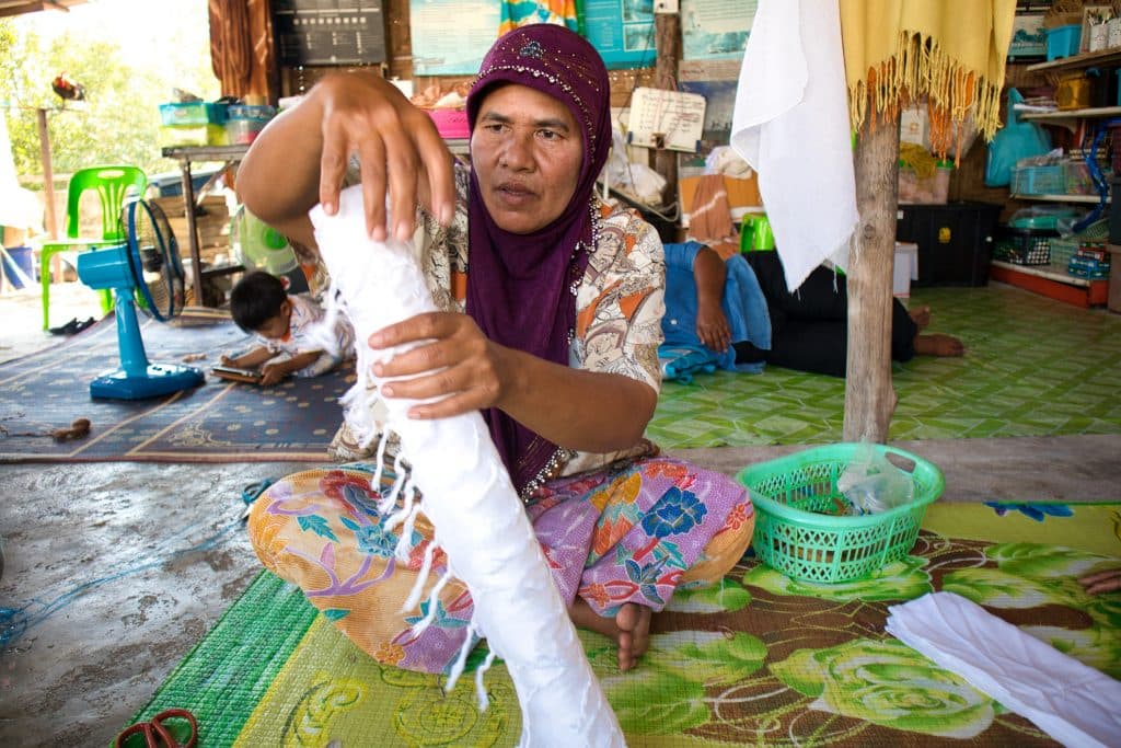 a thai woman sitting on a floor is rolling a white material on a plastic tube. 