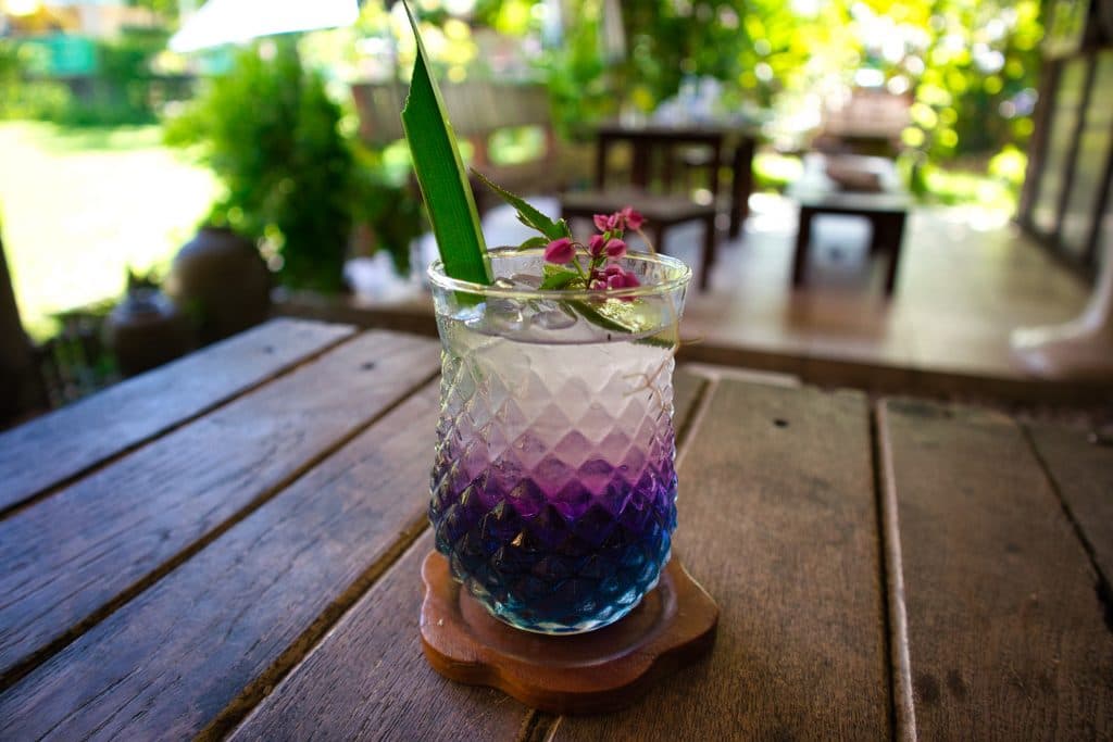 a glass of home made lemonade standing on a table. the water is blud, purpule and white. It has a green leaf and some pink flowers. 
