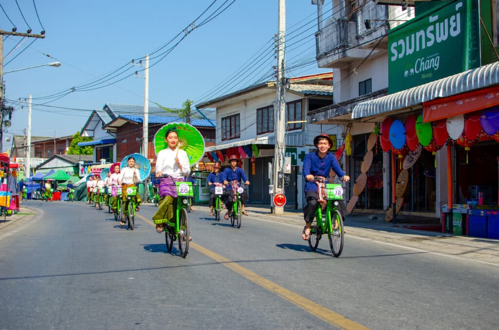A group of ladies dressed in tradtional dresses ride bicycles while holding colourful umbrellas in Bo Sang, Thailand. 
