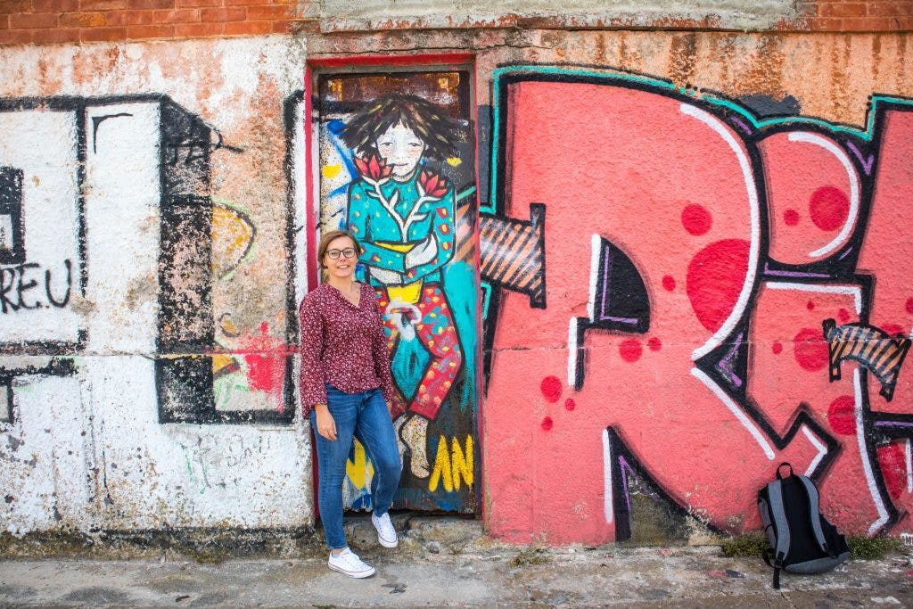 a girl stands at a grafitti wall in almada, lisbon dressed in a purple shirt and jeans smiling