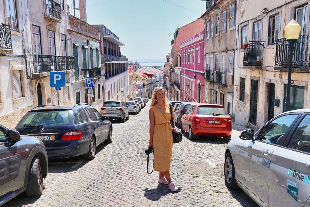 a girl in a yellow dress stands on a street of lisbon with colourful houses and smiles