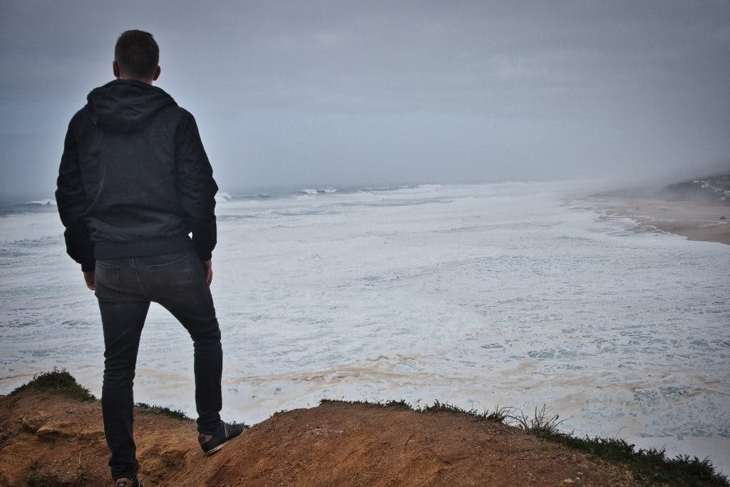 a man dressed in black stands on top of a hill and watches the waves in nazare, portugal