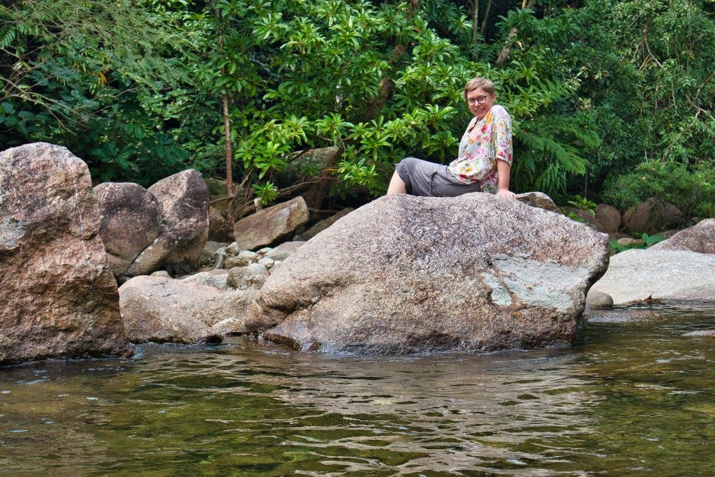 a woman wearing brown trousers and a flowery shirt sits on a rock at promlok waterfall smiling. 