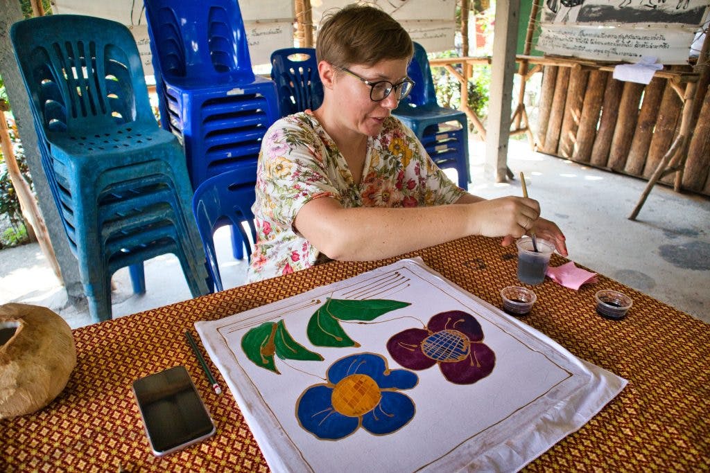a woman dressed in a flowery shirt sits at a table and draws flowers with batik.