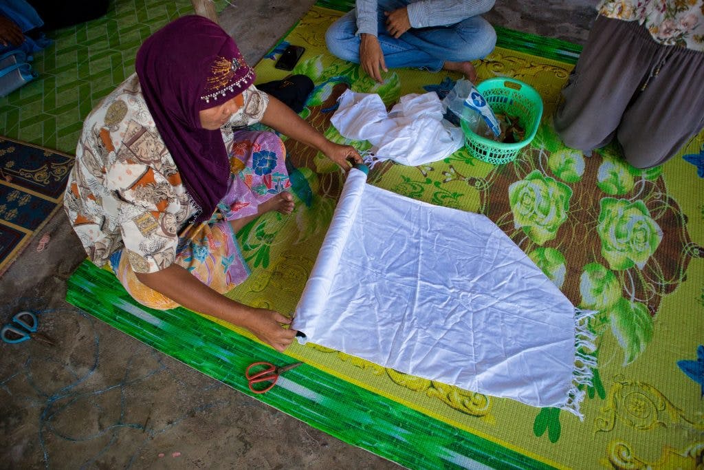 a thai woman is sitting on a floor covered with a yellow matt. she is holding a white material and rolling it on a plastic tube. thailand, krabi, koh klang. 