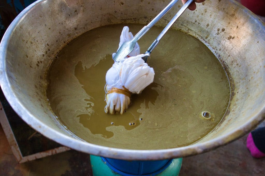 white material held with a metal tool being boiled in green, dirty water in a metal bowl. 