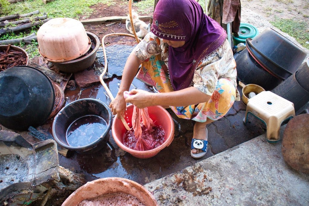 a thai women sitting on the floor washing red material in a red bowl. 