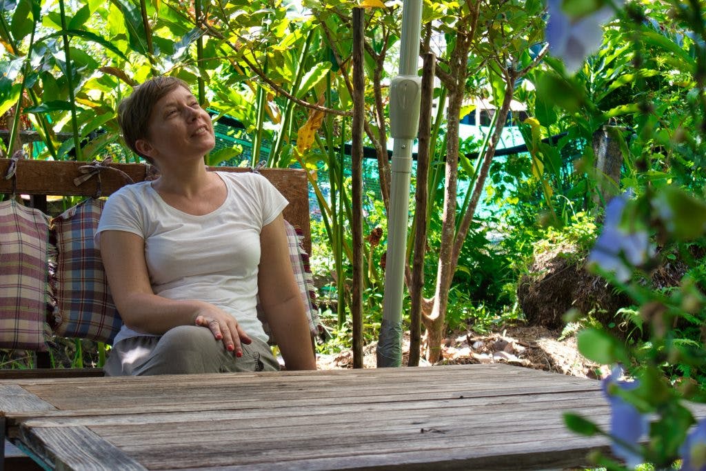 a woman in a white shirt sitting on a wooden bench among green trees. she looks up and smiles a little. 