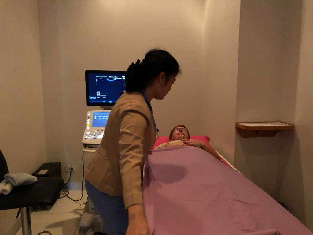 a woman is in bed at ram hospital, chiang mai. a nurse is covering her with a blanket for an ultrasound check.
