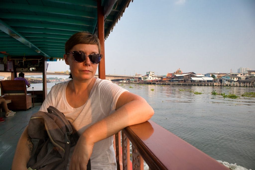 a woman rides a boat from koh kret to bangkok at a sunset. she leans on a rail and looks at the water. 