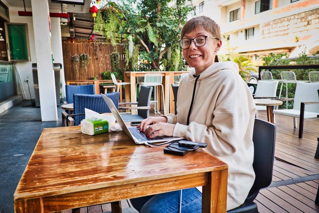 A woman in a white hoodie is sitting at a wooden table with her laptop at smiles at the camera.