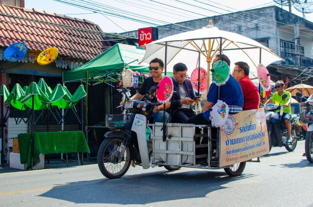A group of men sit in a motorbike cart. The driver is smiling to the camera. During Bo Sang Festival, Chiang Mai. 