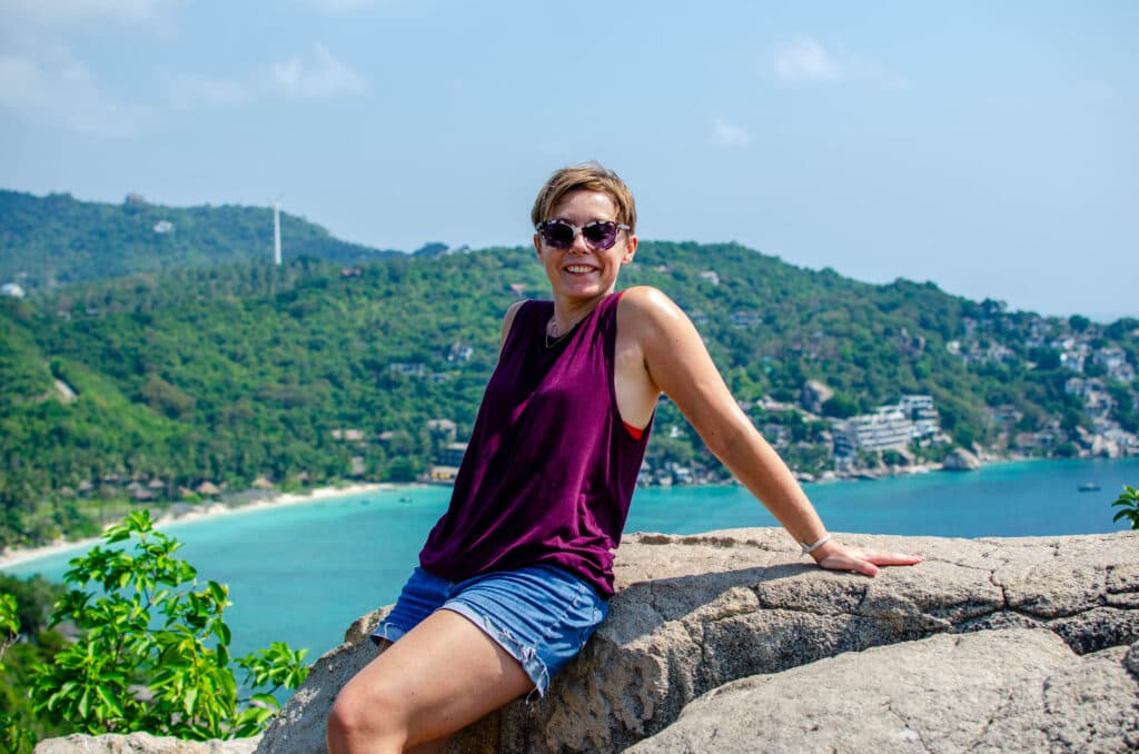 A woman sitting on a rock on the viewpoint on koh tao. She is smiling at the camera. Behind her there is a beach and a forest. The view overlooks the bay.