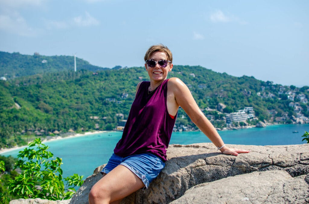 A woman sitting on a rock on the viewpoint on koh tao. She is smiling at the camera. Behind her there is a beach and a forest. The view overlooks the bay. 