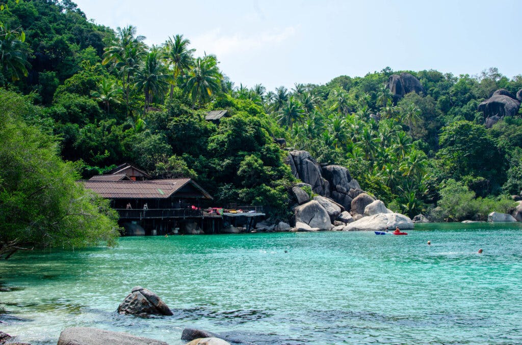 Crystal clear waters surrounding the bay on koh tao. in the distance tehre are poeple kayaking and a wooden house with a view terrace. 