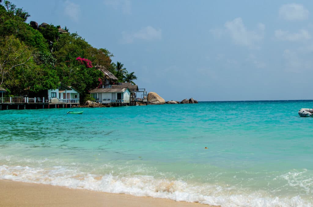 Blue waters on Shark Bay, Koh Tao, thailand. In the distance we see a couple of houses and a hill with forest.