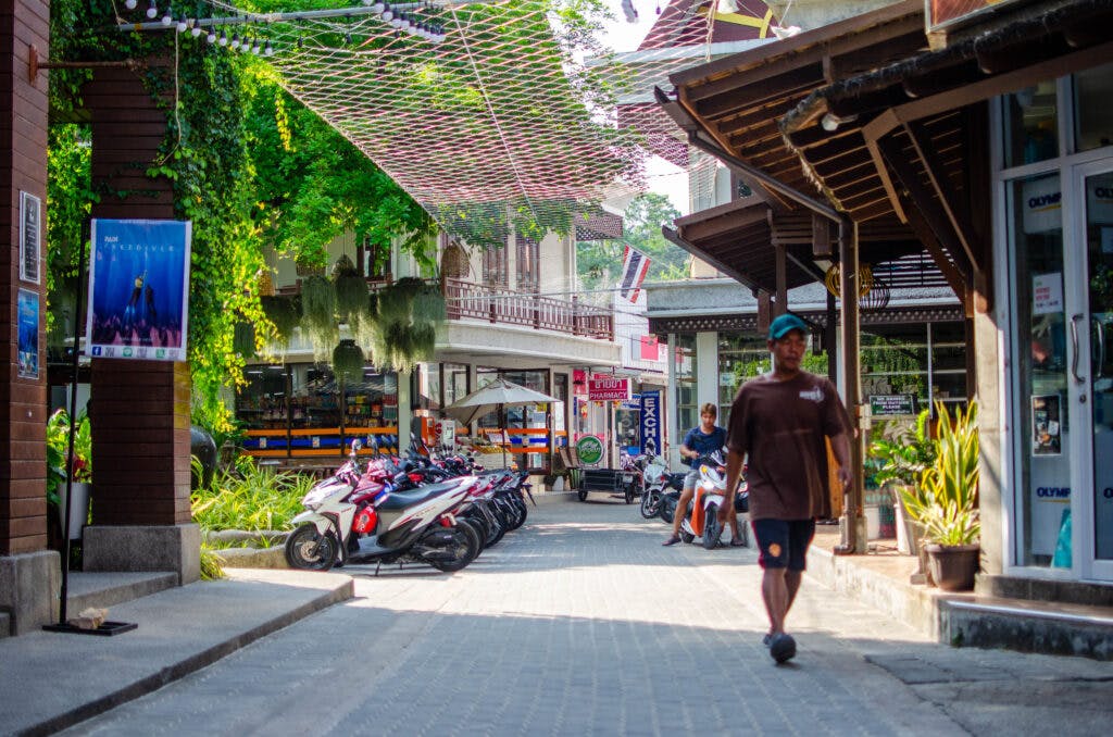 Główna ulica na Koh Tao w Tajlandii. 7-11 widziany z daleko, skutery stoją na ulicy, chodnikiem przechodzi się mężczyzna. 