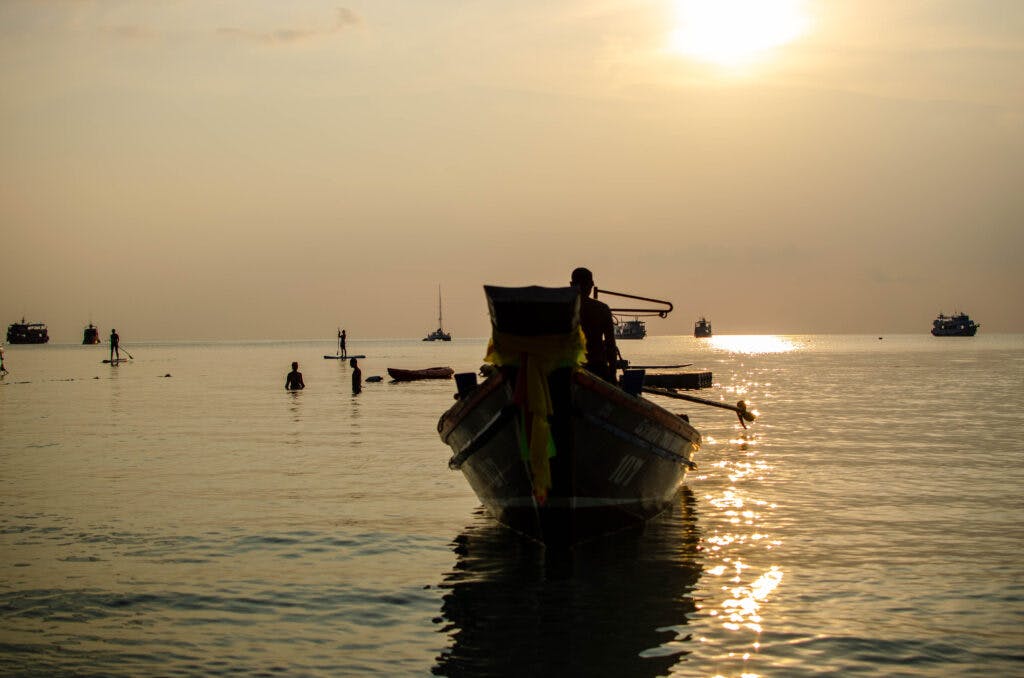 Zachód słońca na Koh Tao. Parę osób pływa na supach, mężczyzna siedzi w łódce, w dali widać statki. 
