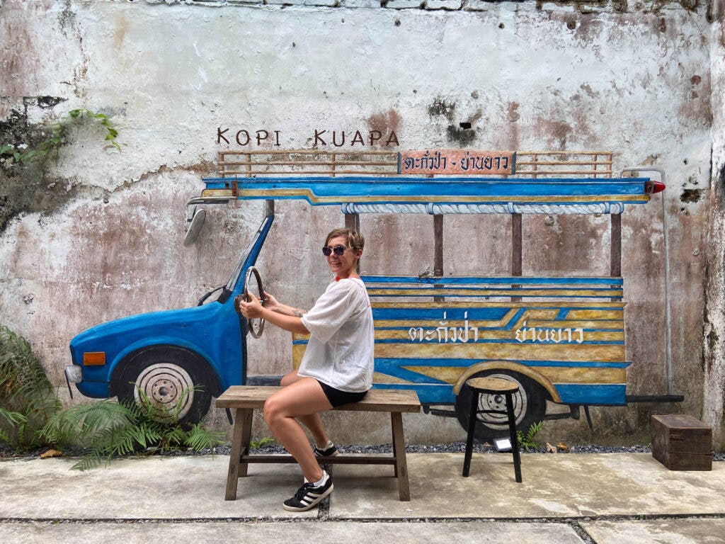 A woman sitting on the bench in front of a grafitti of a car in Thailand. She looks like she's driving it. 