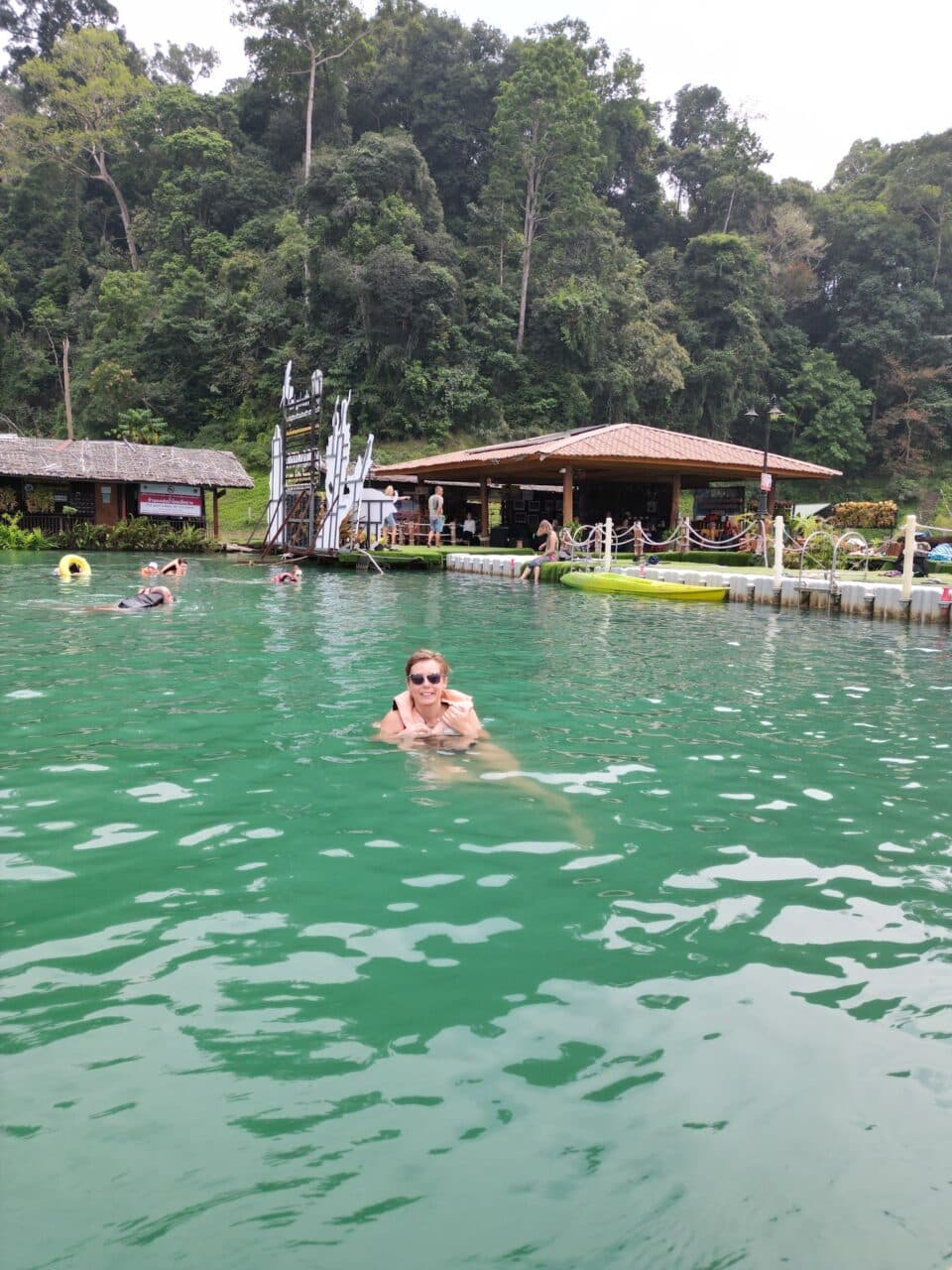 Joanna swims in the khao sok lake, behind her there is a small hut on the water.