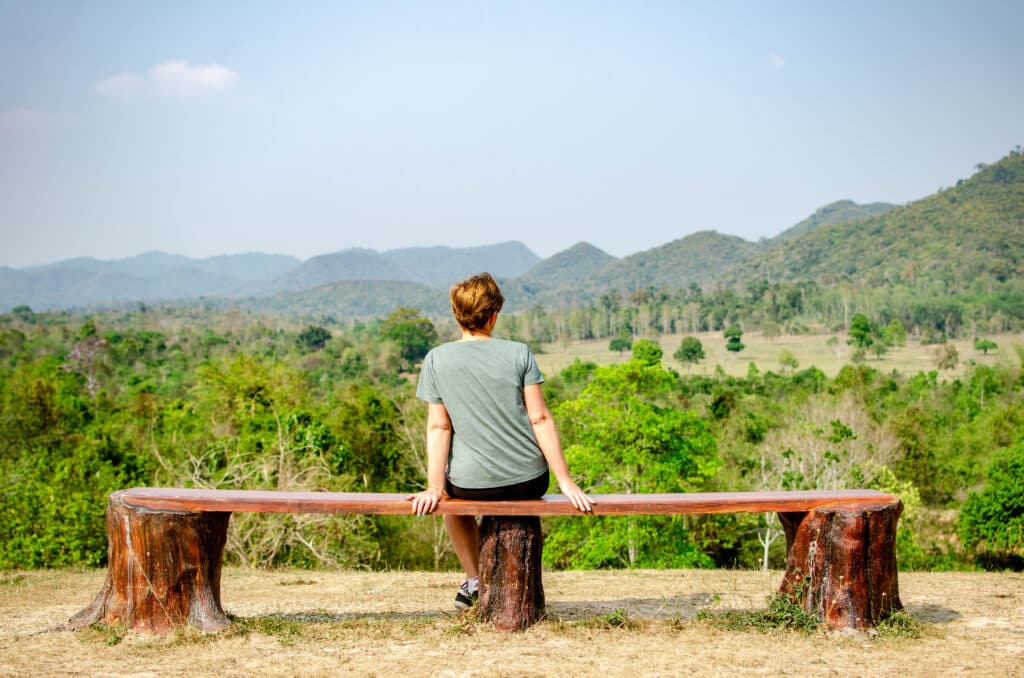 A woman sitting with her back to the camera. She looks at the fields and is waiting for the wild elephants to emerge, national park, thailand.