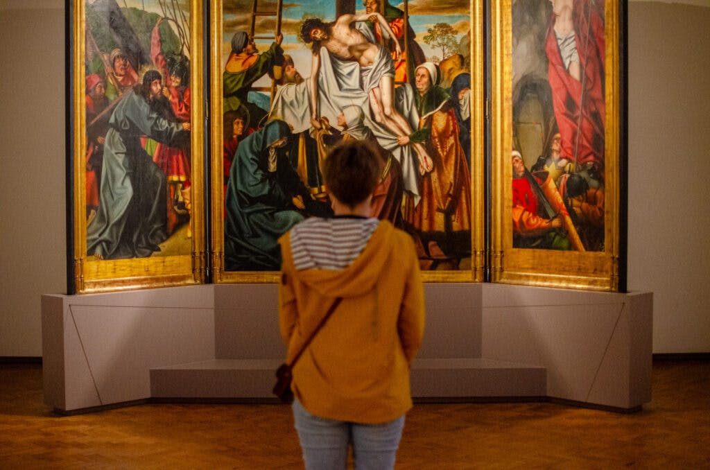 Kobieta w żółtej kurtce w muzeum w Lizbonie ogląda duży obraz przedstawiający Jezusa ściągniętego z krzyża. 