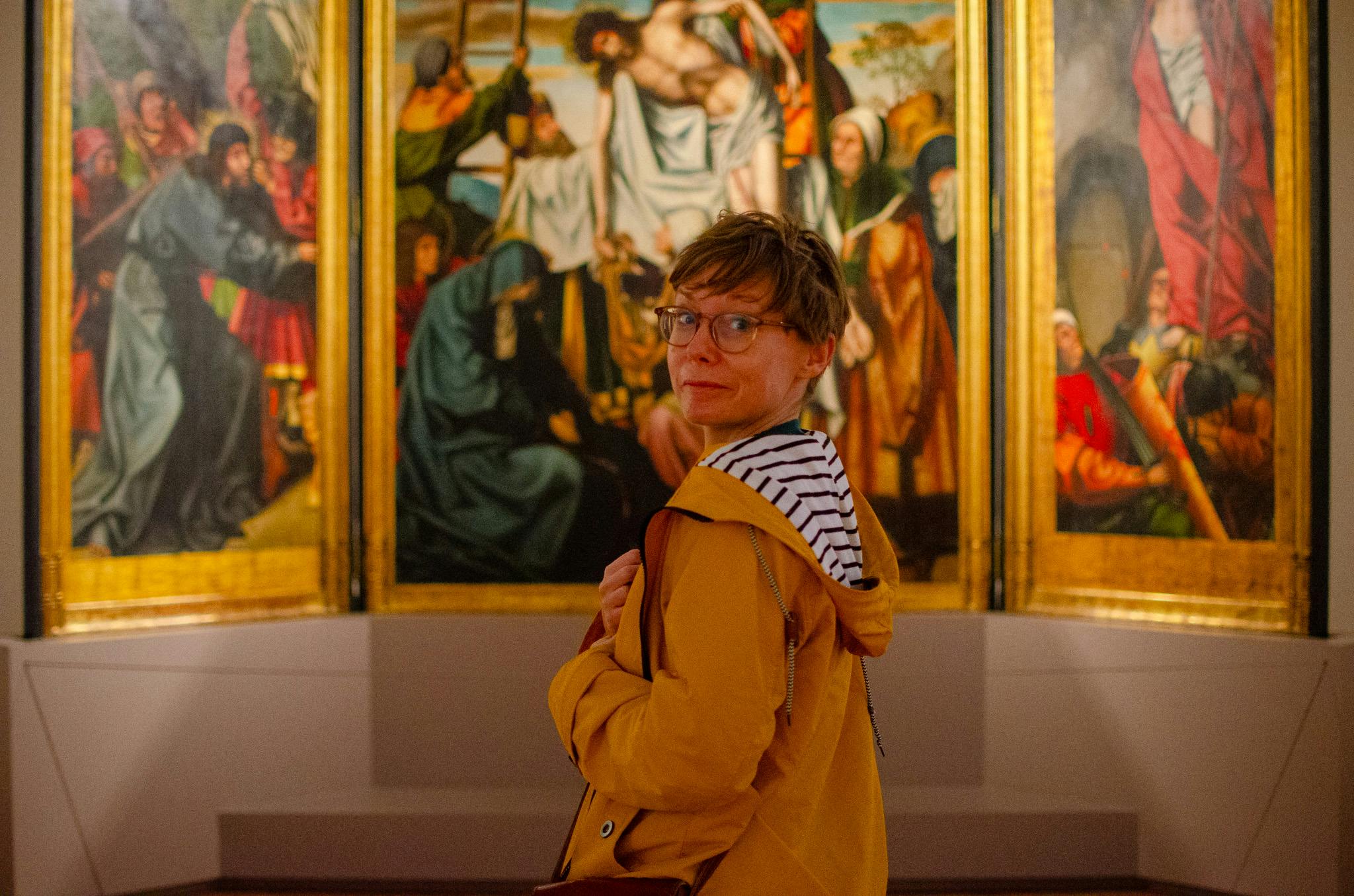 Kobieta na tle obrazu patrzy się w kamerę. Ma na sobie żółtą kurtkę. Muzeum Sztuki Starożytnej.