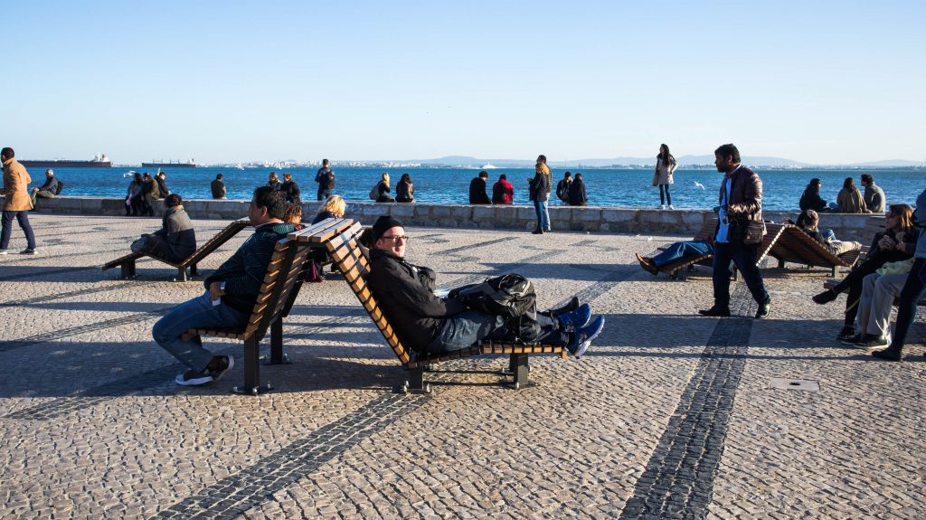 people resting on benches on a sunny day by the river in lisbon