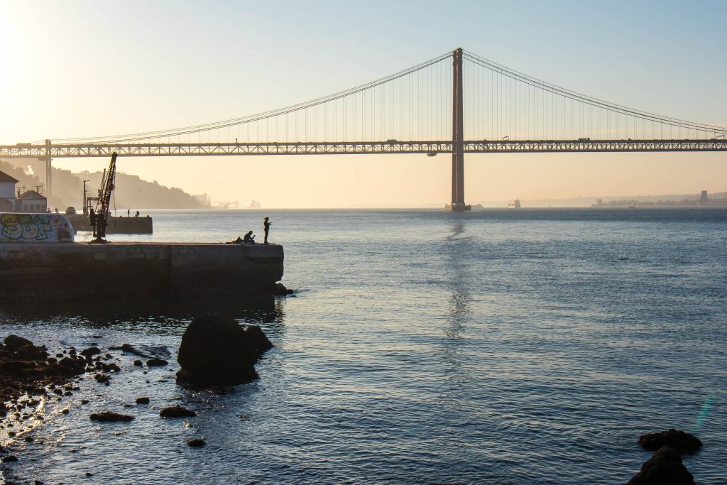 a fisherman stands on the shore of the tagus river with the 25th april bridge in the background at the sunset