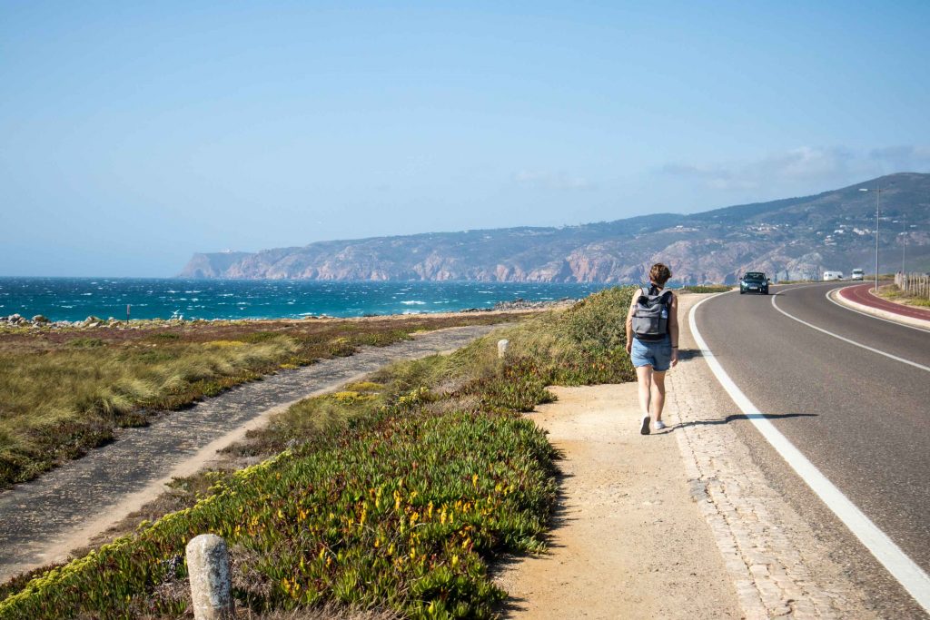 a girl walks from cascais to guincho by the side of the road with the ocean on her left.