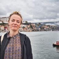 a girl stands at the river in porto smiling lightly on a cloudy day