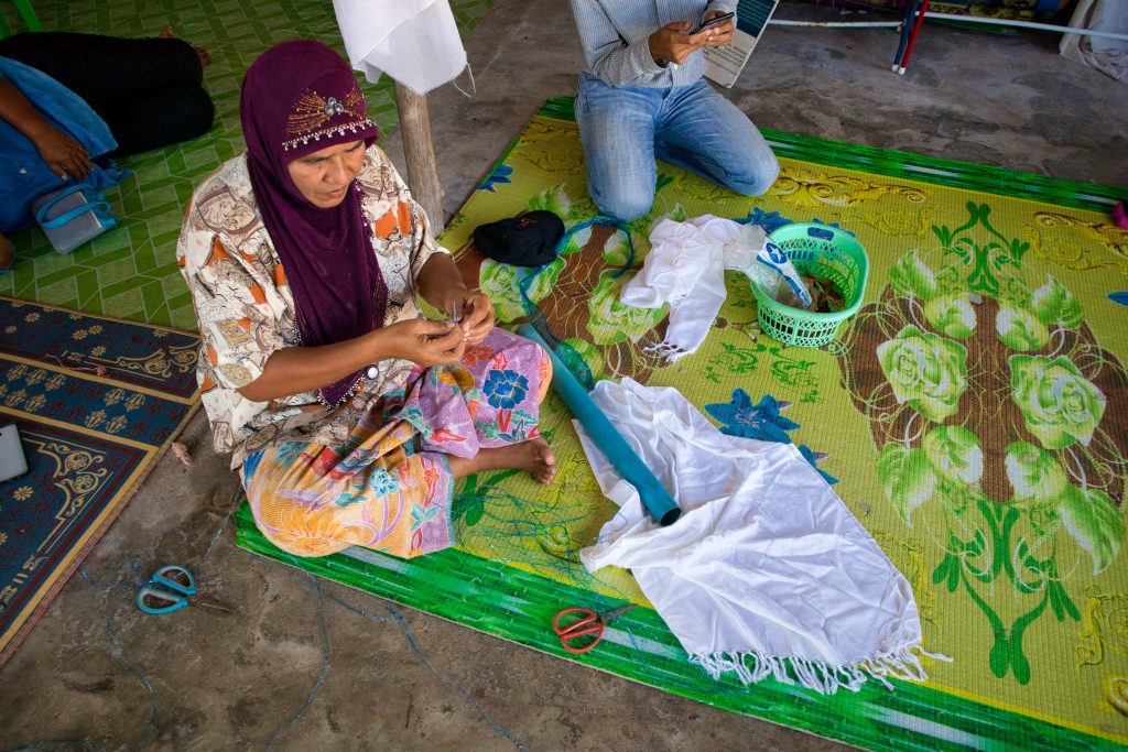 a thai woman sits on a yellow, flowery matt, on the floor. she is folding a material. she is wearing a red scarf and a flowery blouse. 