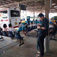 a man standing at a station in nakhon si thammarat listening to music while other passengers are waiting