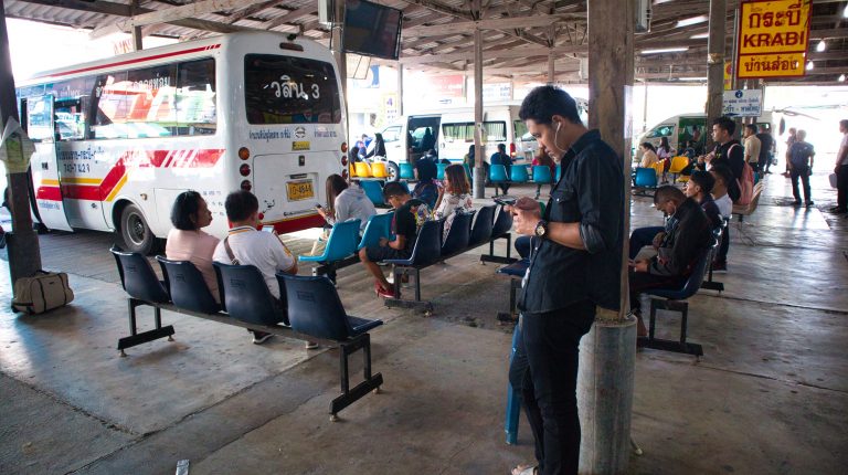 a man standing at a station in nakhon si thammarat listening to music while other passengers are waiting