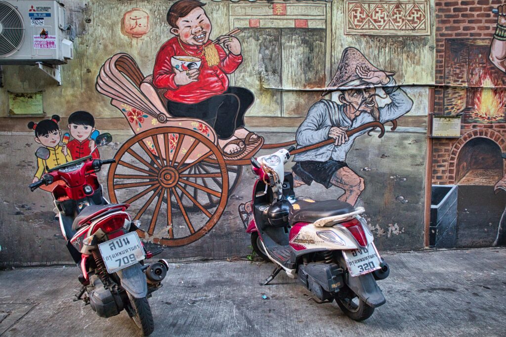 two motorbikes standing in talad noi in front of a grafitti, showing an old man pulling a cart with a fat chinese man eating noodles. 