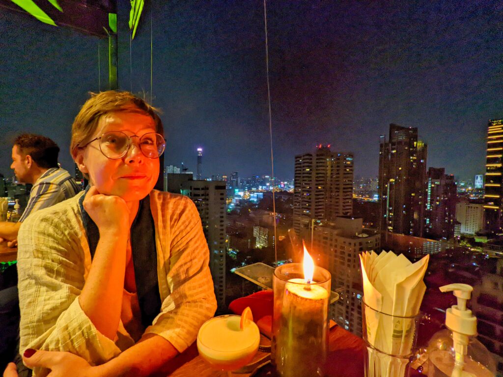 Rooftop w Bangkoku. Dziewczyna siedzi przy drinku i stoliku z widokiem na bangkok w nocy.